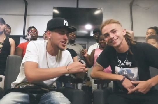Two men are sitting next to each other and one has a ny hat on