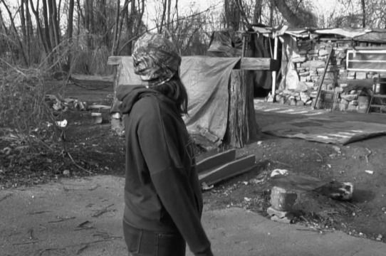 A black and white photo of a person standing in front of a shack.