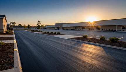 Sunset over an empty parking lot beside low industrial buildings, with a long dark road in the foreground