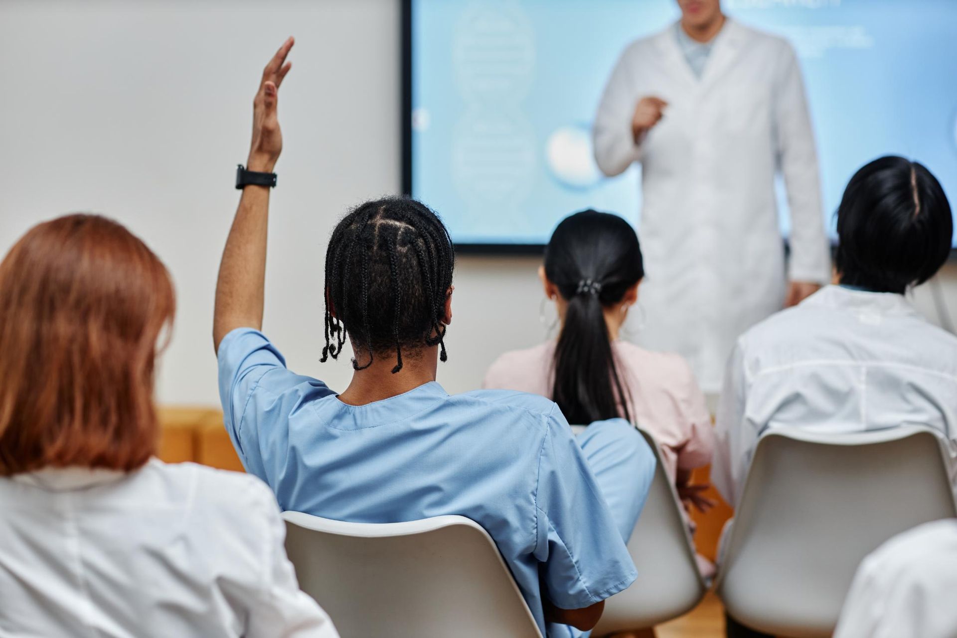 Audience raising hands in a classroom or seminar, facing a presenter in white.