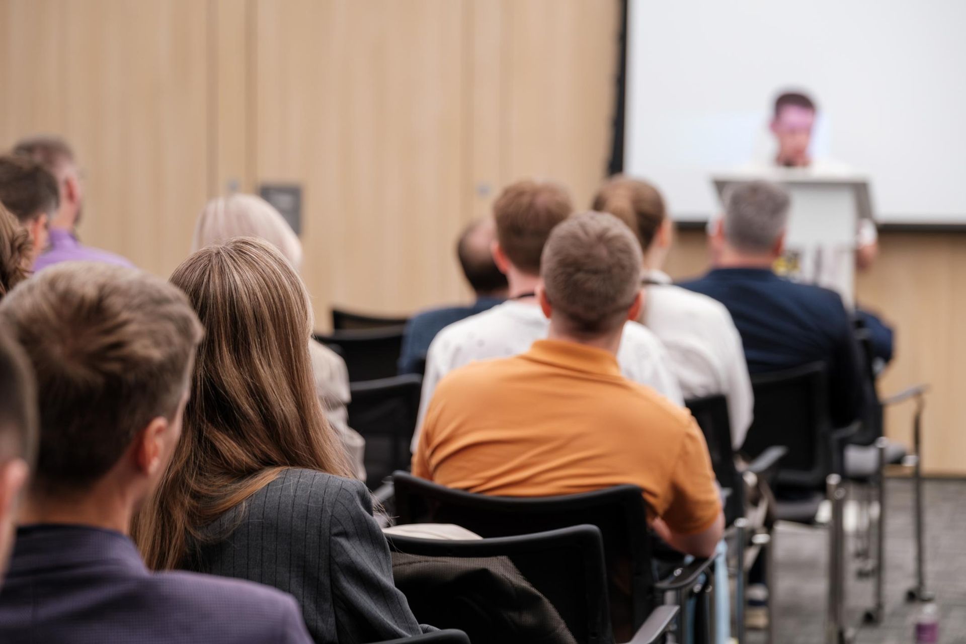 Audience seated in a conference room, listening to a presentation at the front.