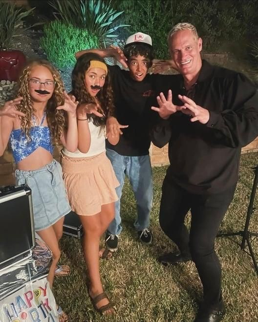 Four costumed people posing outdoors at a nighttime party, smiling beside colorful lights.