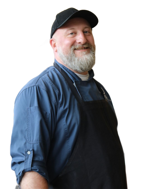 A smiling person with a beard, wearing a black cap, blue shirt, and black apron.