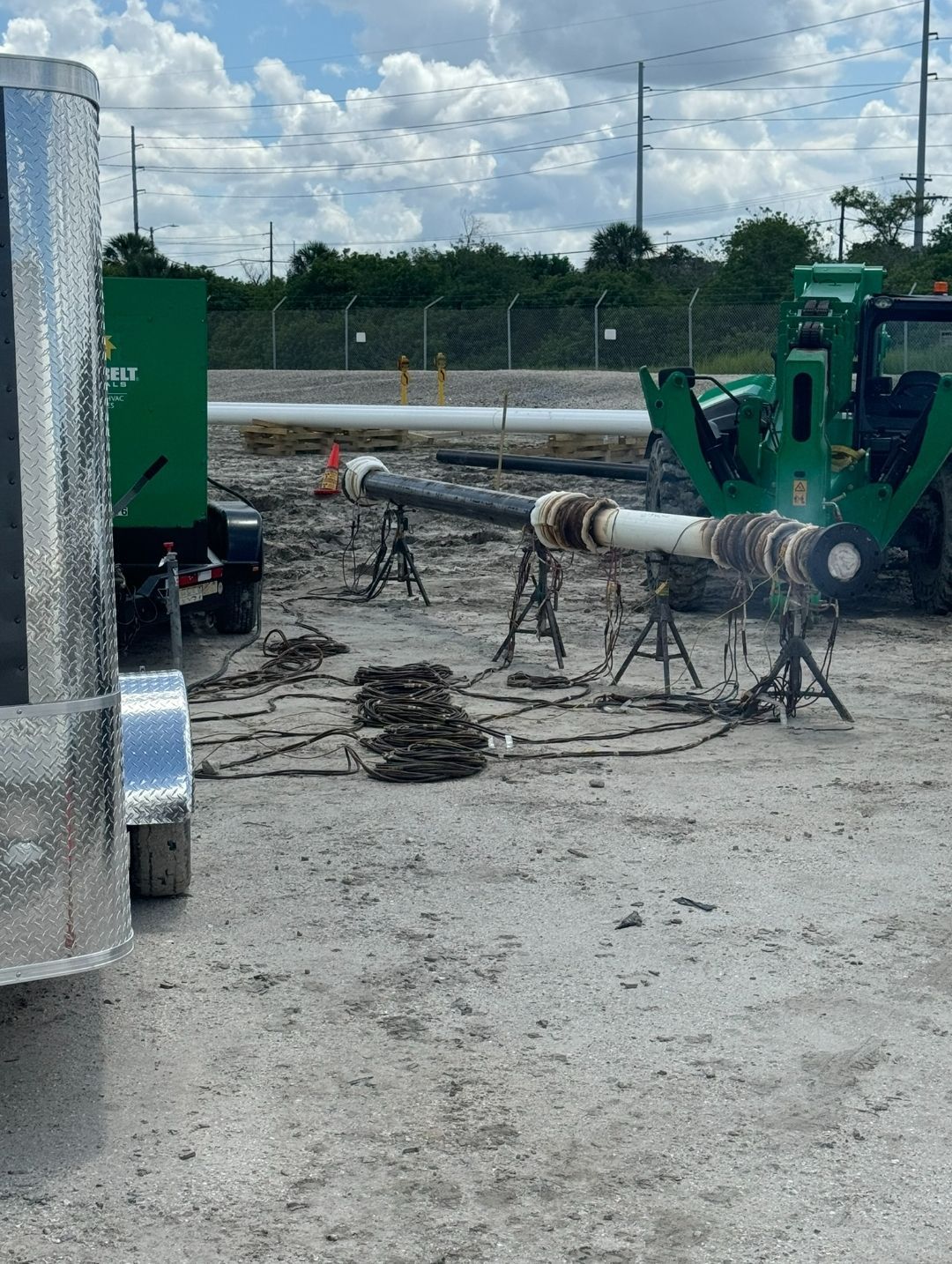Industrial site with green machinery, a horizontal pipe on supports, and coiled cables on gravel.