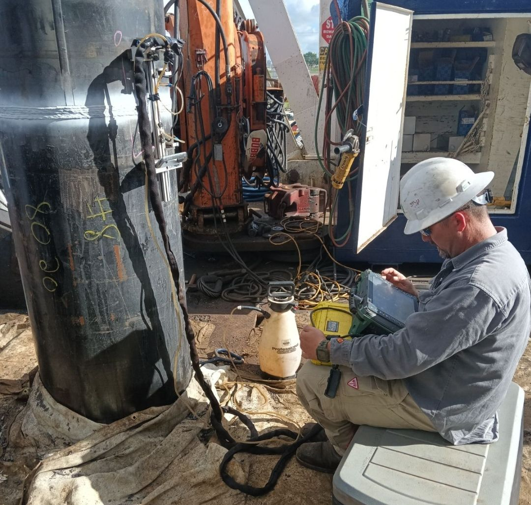Worker in a white hard hat using a handheld device beside industrial equipment and cables outdoors.