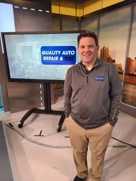 Smiling man stands beside a TV showing “Quality Auto Repair & Tire” in a studio setting | Quality Auto Repair Tire & Auto Sales