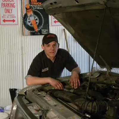 Mechanic working under a raised car hood in a garage, with tools and safety signs in the background | Quality Auto Repair Tire & Auto Sales