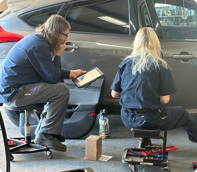 Two people seated on low stools sketching by a parked gray car outdoors | Quality Auto Repair Tire & Auto Sales