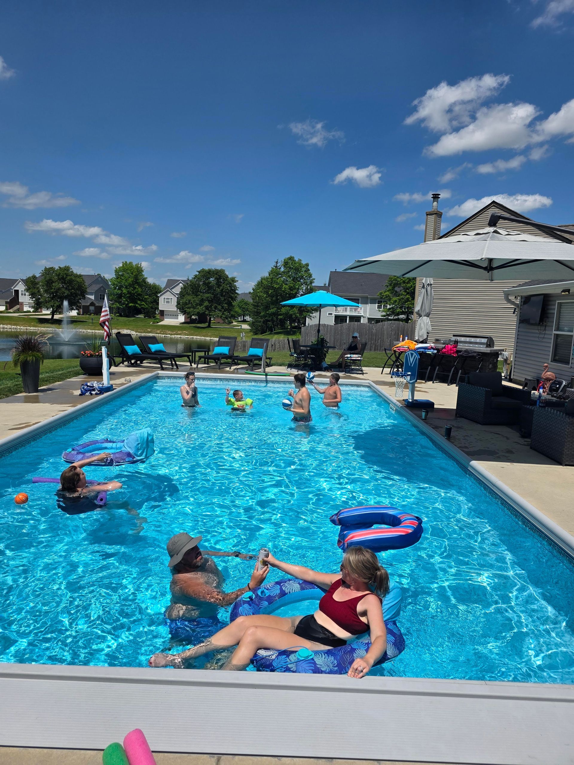 People in a pool on a sunny day. Some are on floats. Blue water, clear sky, and a backyard setting.