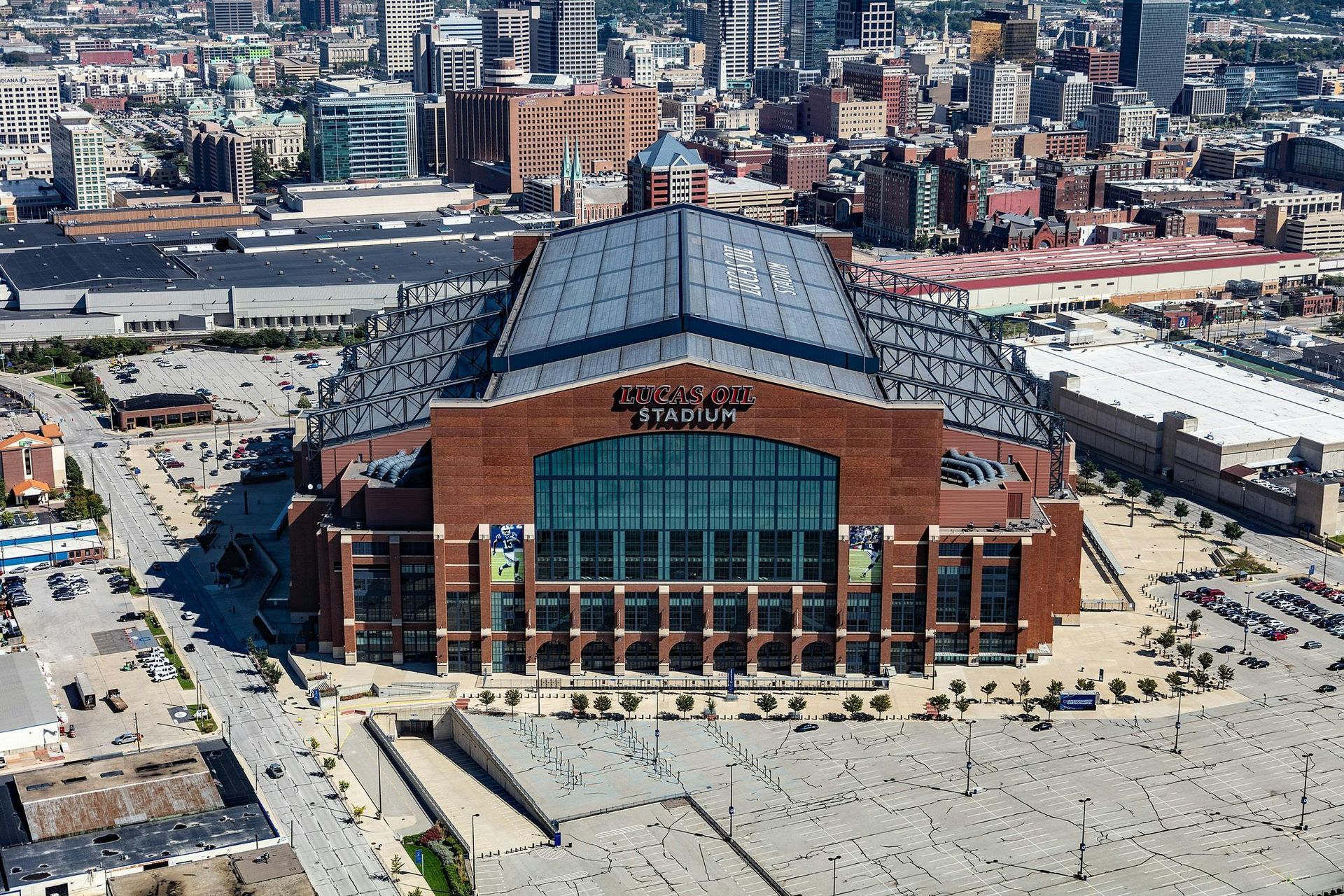 Lucas Oil Stadium, Indianapolis, Indiana. Large brick stadium with glass windows and a metallic roof, surrounded by city buildings.