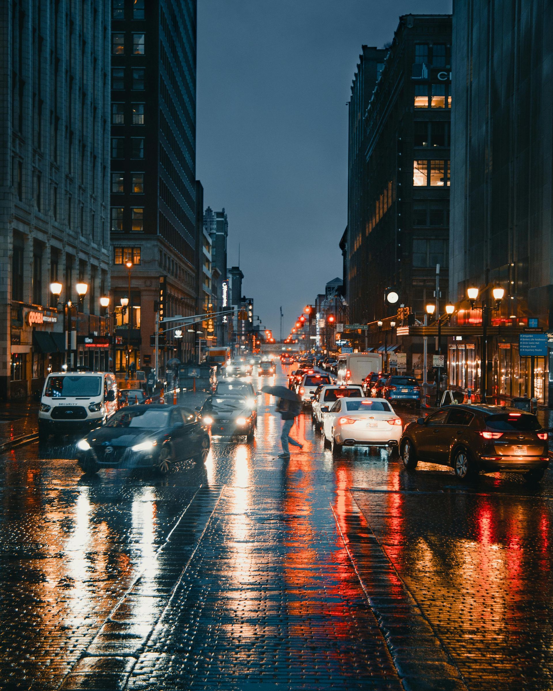 Rainy city street at night, with cars and buildings reflecting lights on the wet pavement.