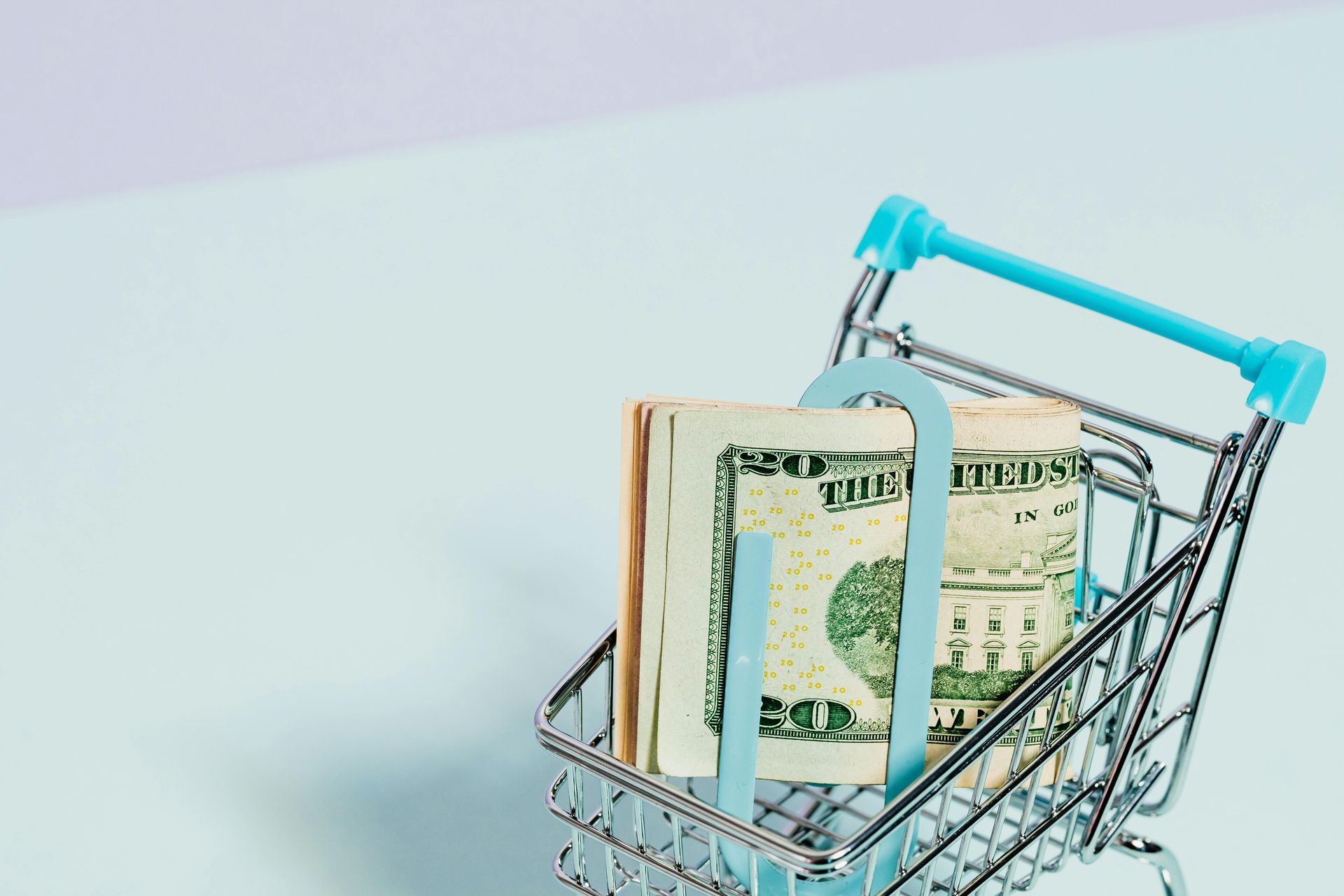 Miniature shopping cart with a stack of U.S. dollar bills, secured by a blue binder clip, on a light blue surface.
