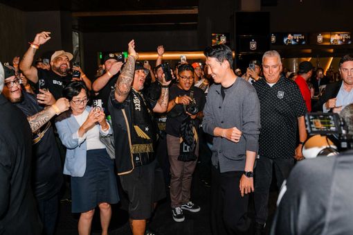 Crowd cheering and filming a man in a gray shirt walking through an indoor event venue