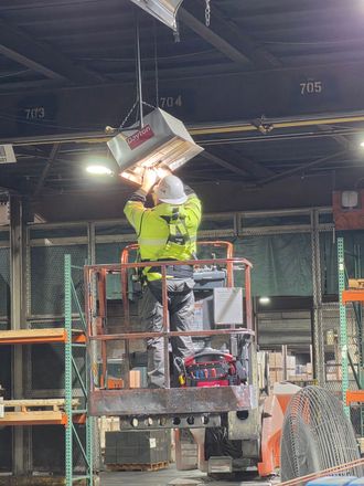 Person standing at an outdoor service counter, reaching into a metal cabinet with a backpack sprayer on
