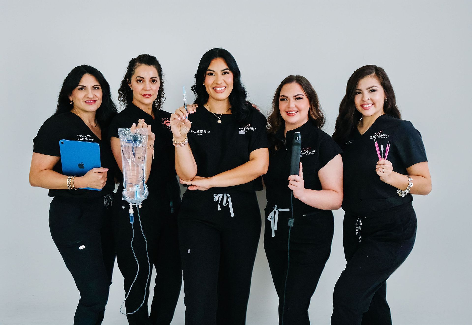 Five women in black scrubs, holding medical tools, smiling against a white backdrop.