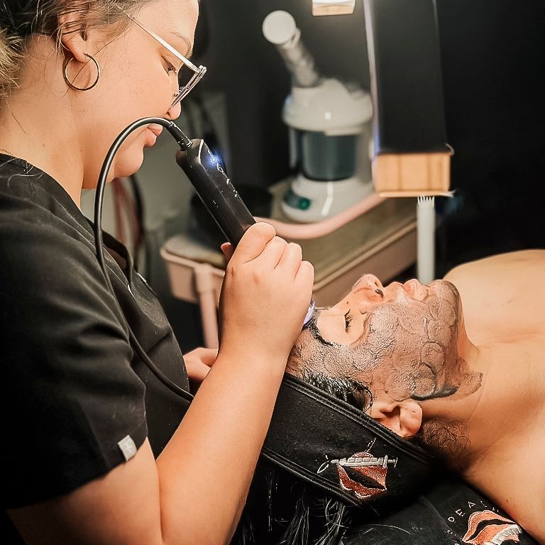 Woman performs facial treatment on a man. She holds a device to his face; he lies back.