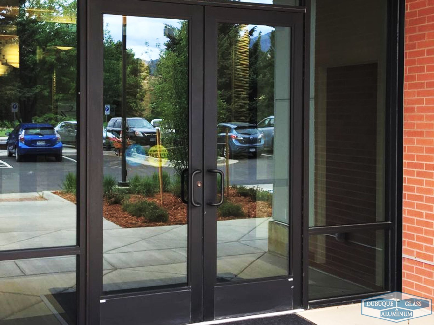 Glass double doors at a building entrance with reflections of parked cars and trees outside