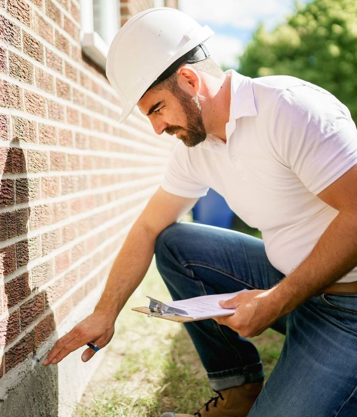 A man wearing a hard hat is kneeling down and looking at a brick wall.