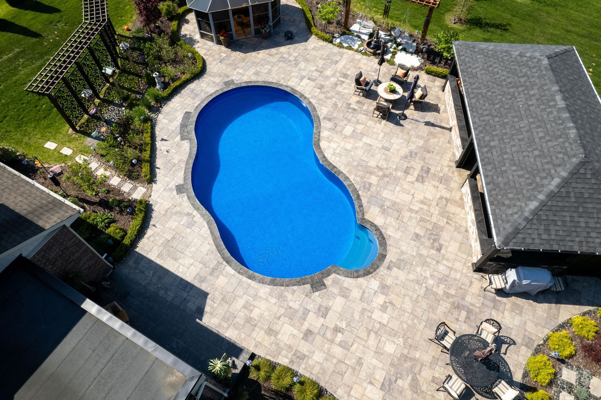 Aerial view of a bright blue, kidney-shaped swimming pool surrounded by a large stone patio, gazebo, and pergola.