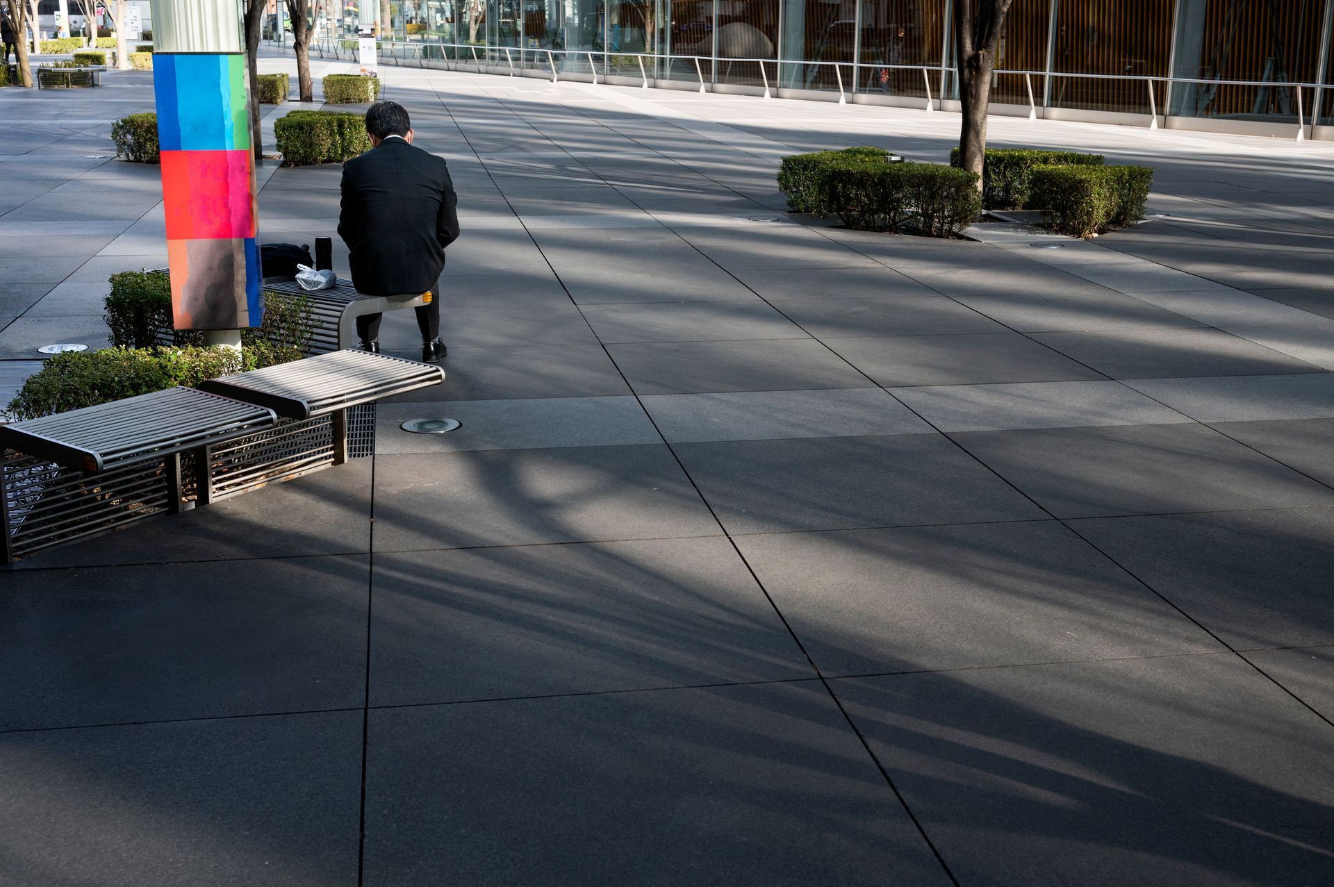 A person sits on a bench in a city square. A colorful column and two benches are in view.