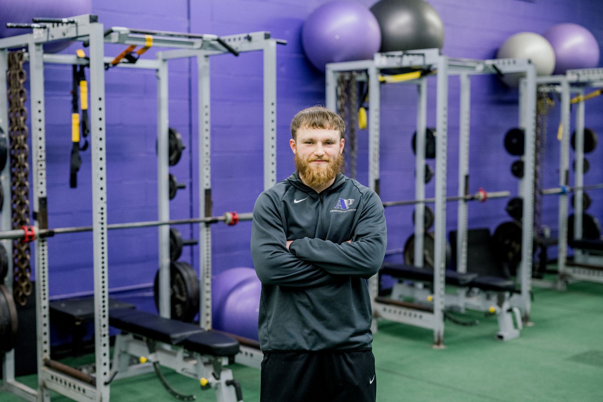 Man standing with arms crossed in a gym, with exercise racks and purple stability balls behind him.