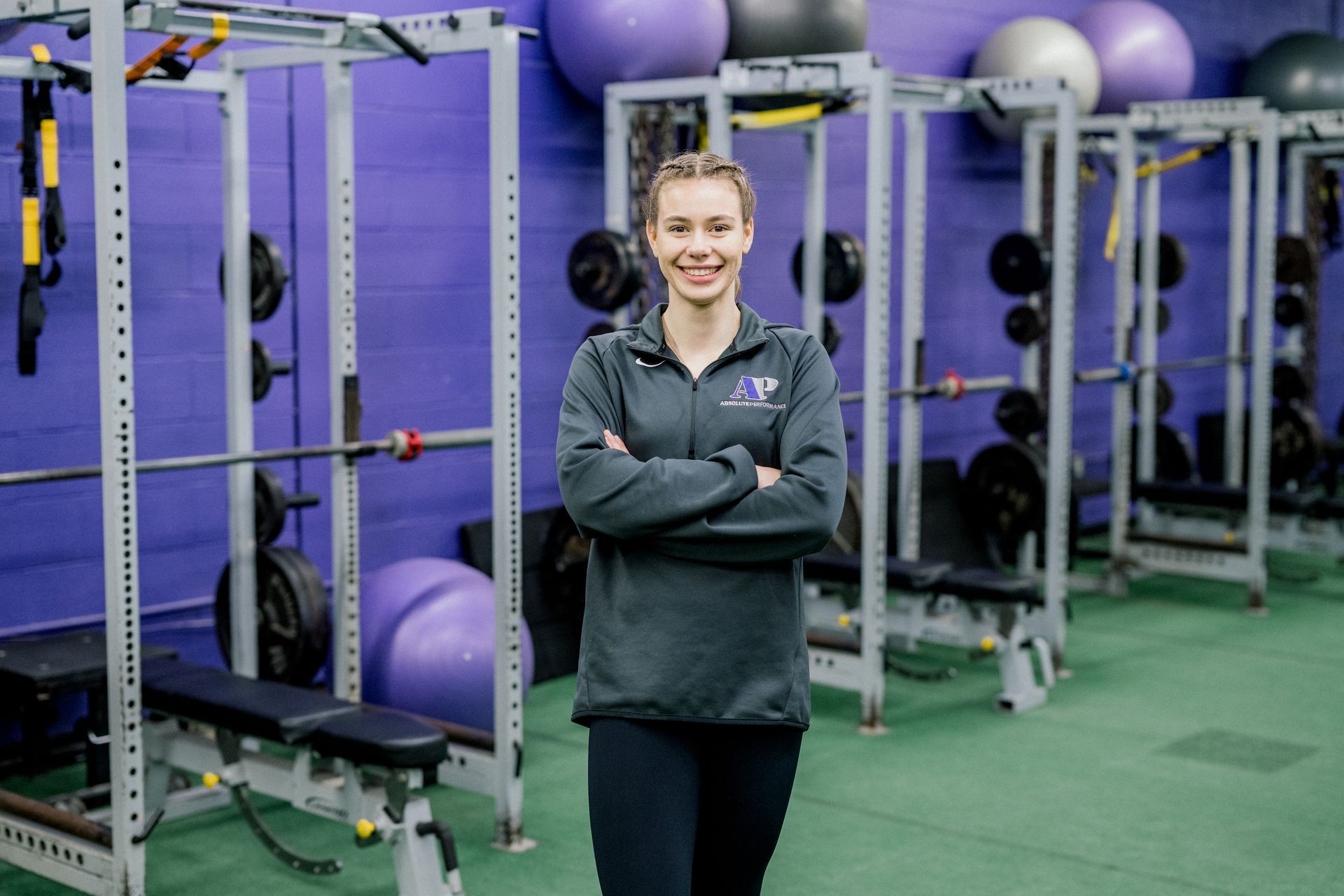 Woman standing in a gym, arms crossed, with purple exercise equipment and exercise balls behind her