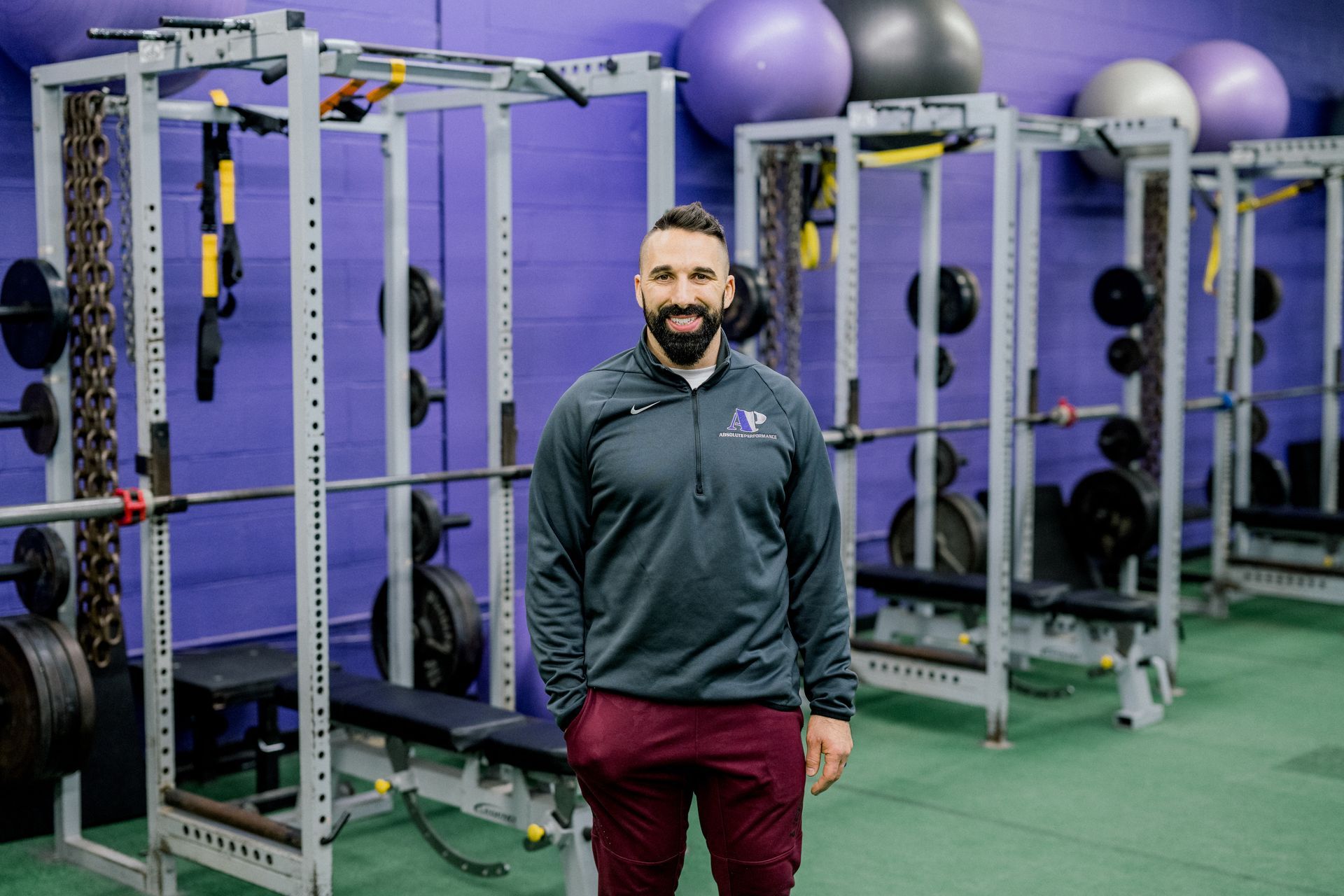 Man standing in a gym with cable machines and exercise balls against a purple wall.