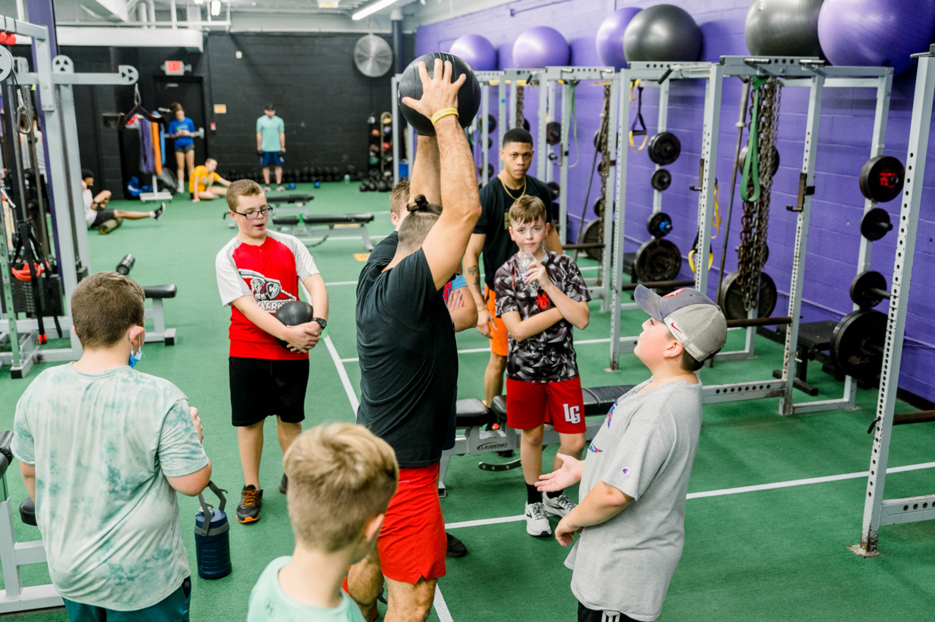 Group exercising in a gym, with one person lifting arms overhead among others on green turf.