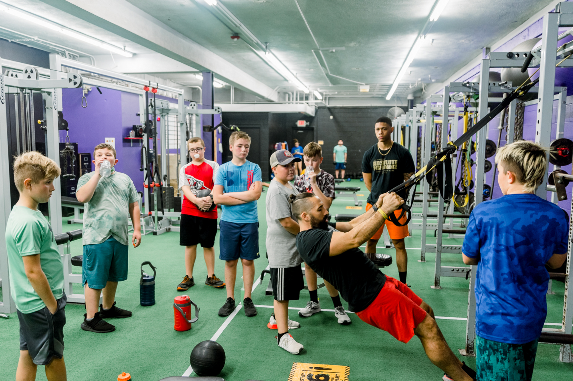 People exercising in a gym, with one person suspended horizontally while others watch nearby.