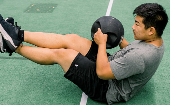 Man holding a medicine ball while assisting a seated athlete doing core exercises on an indoor turf field