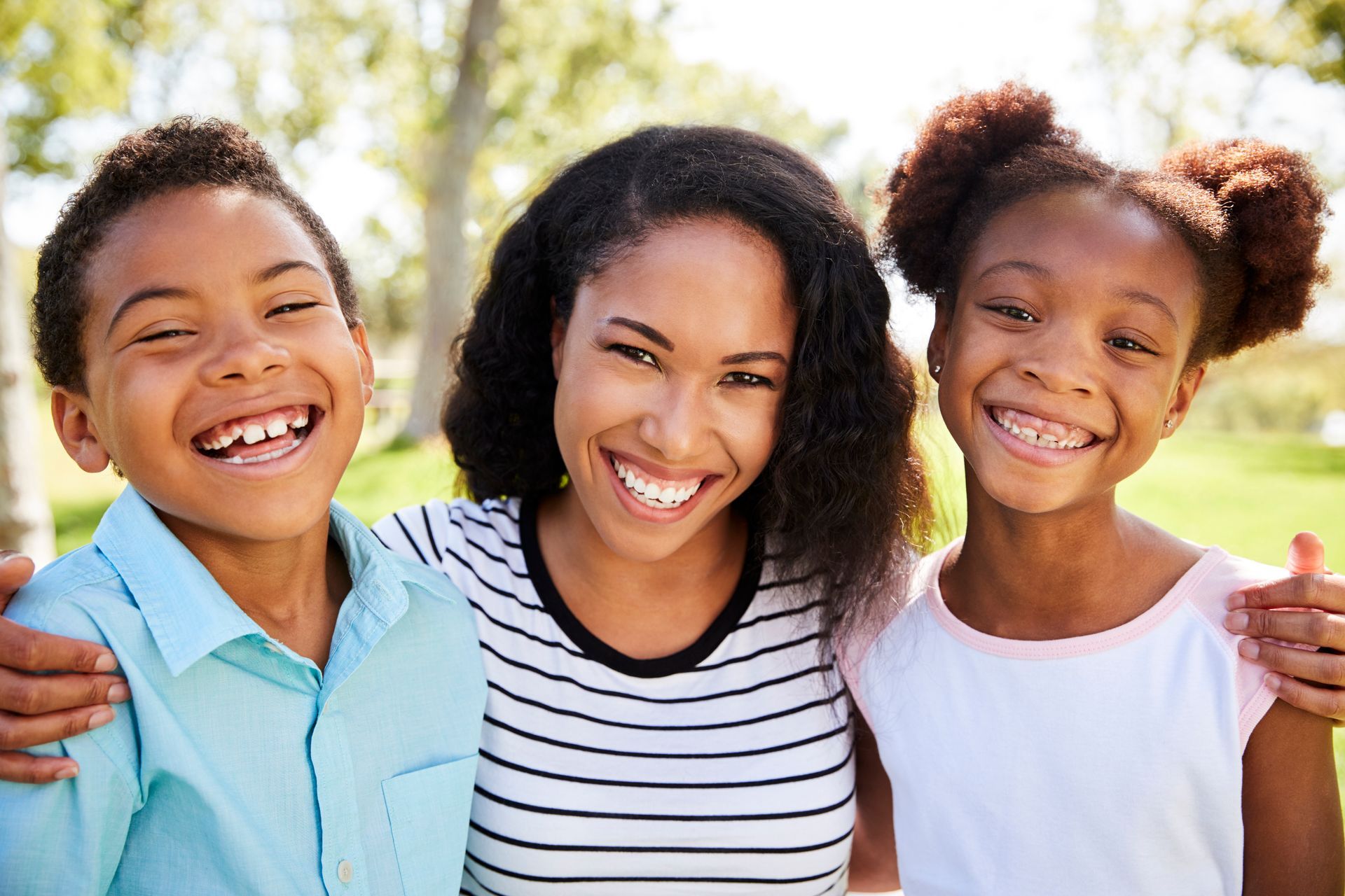 Woman with two smiling children, outdoors, embracing, sunny day.