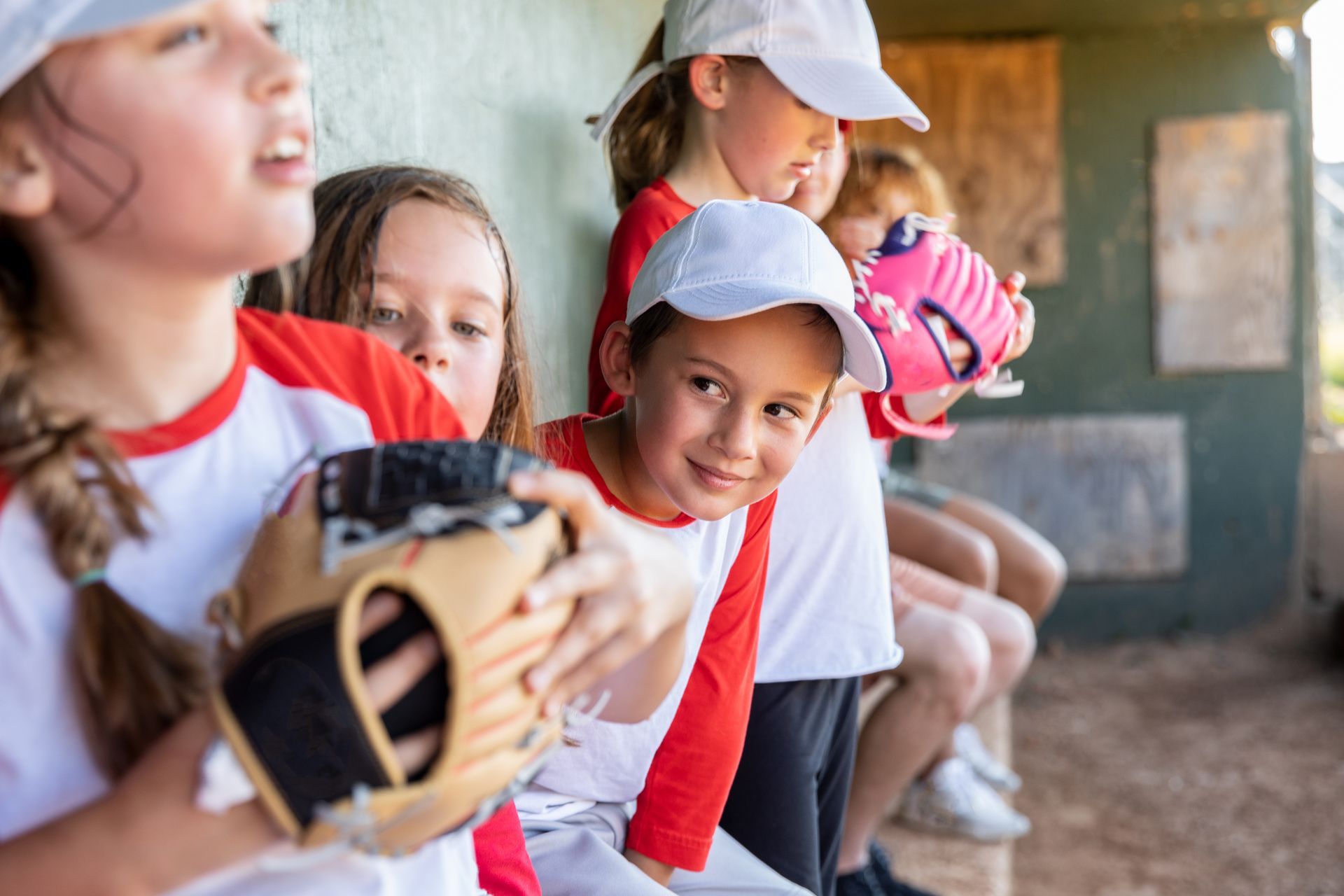 Children in baseball uniforms in a dugout, some holding gloves, smiling.