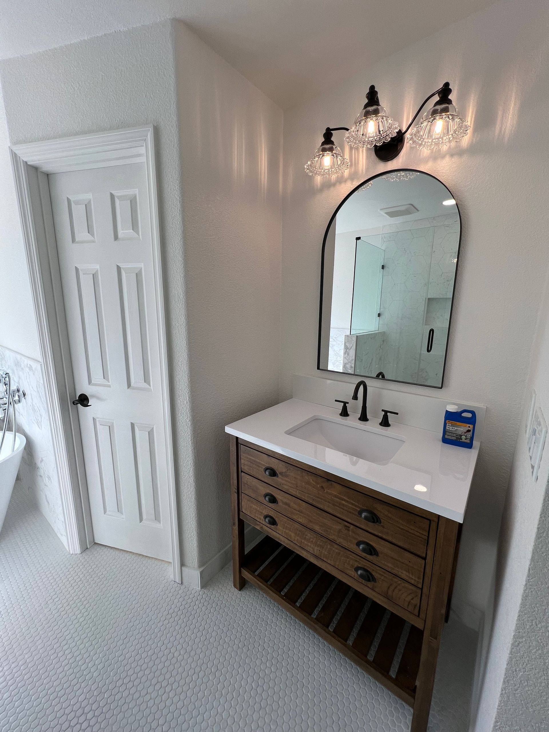 Bathroom with white penny tile, a wooden vanity with an arched mirror, and a three-light fixture.
