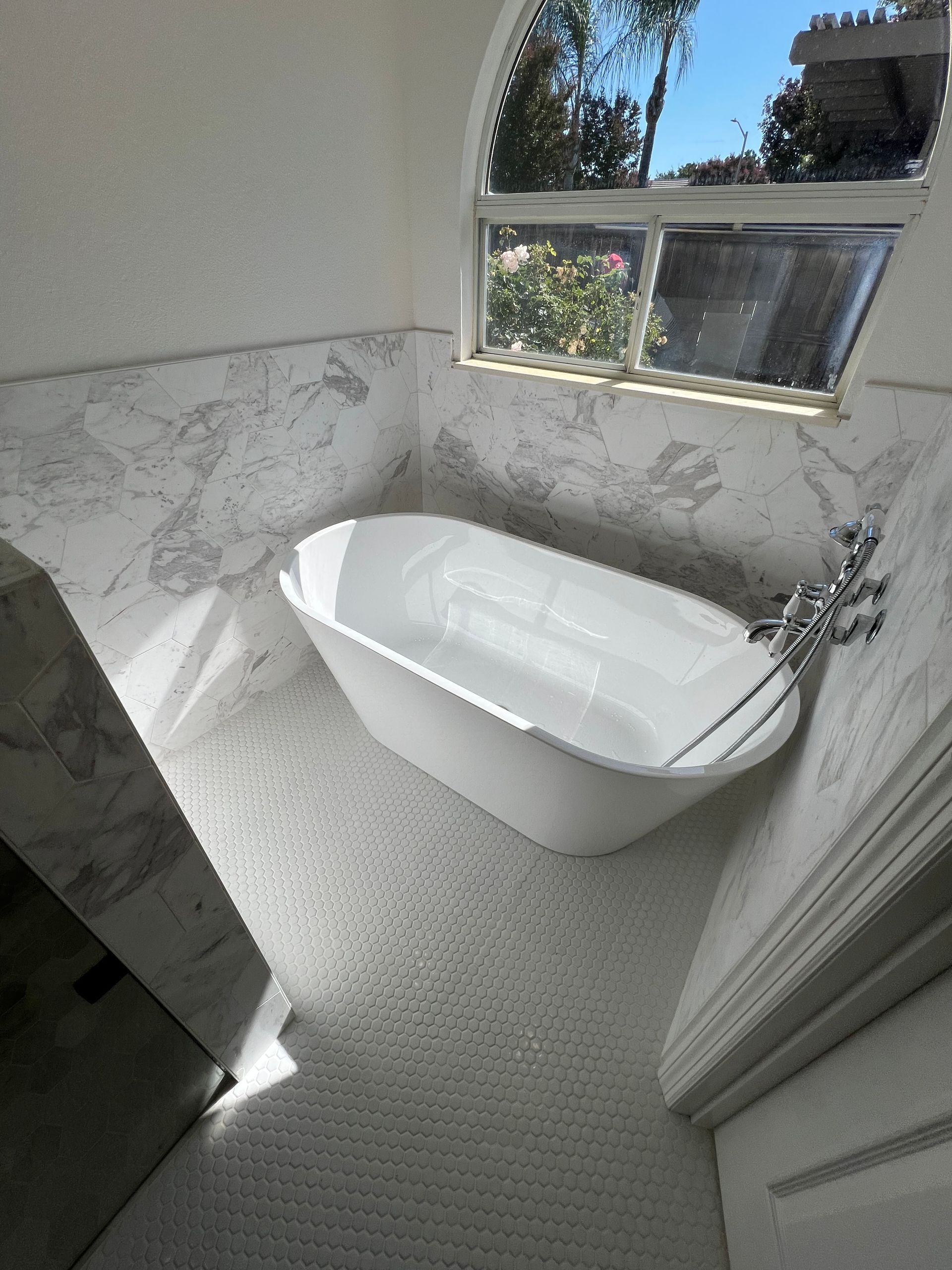 White bathtub in a bathroom with marble walls and a window. Sunlight streams in.