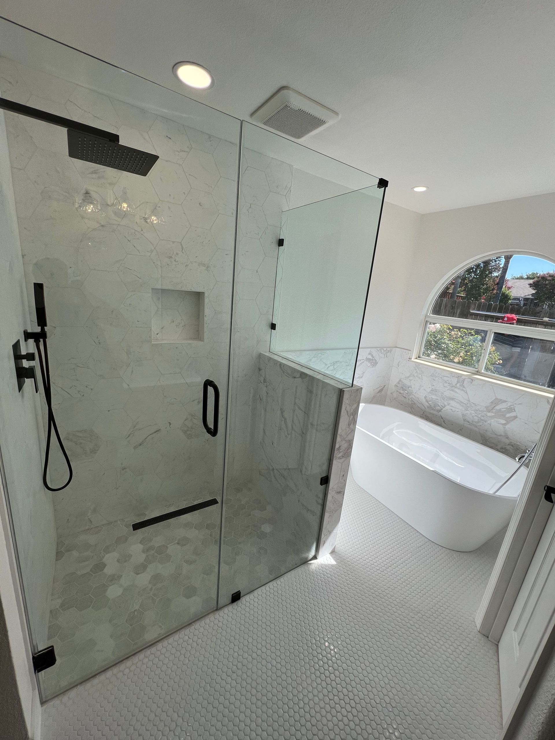 A modern bathroom features a glass shower, white tile, and a freestanding tub near an arched window.