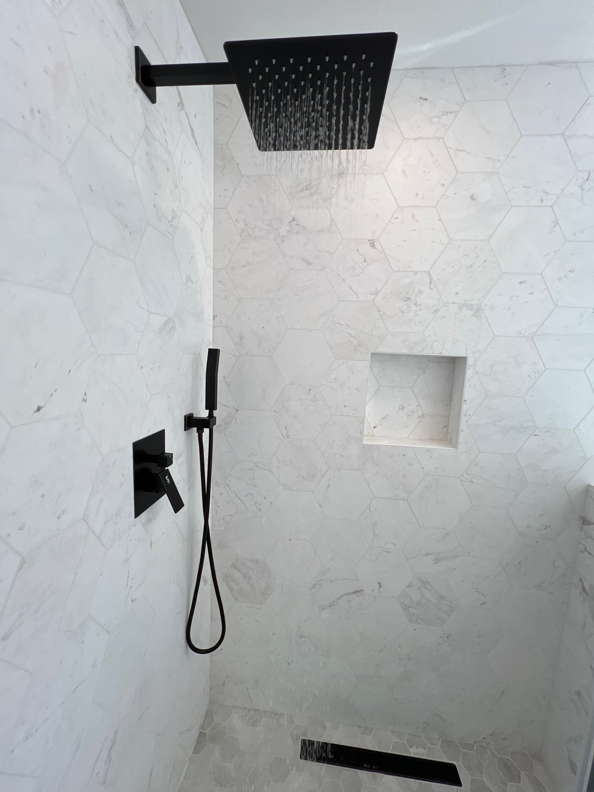 A modern shower with black fixtures against white marble tile. The showerhead is on, dispensing water.