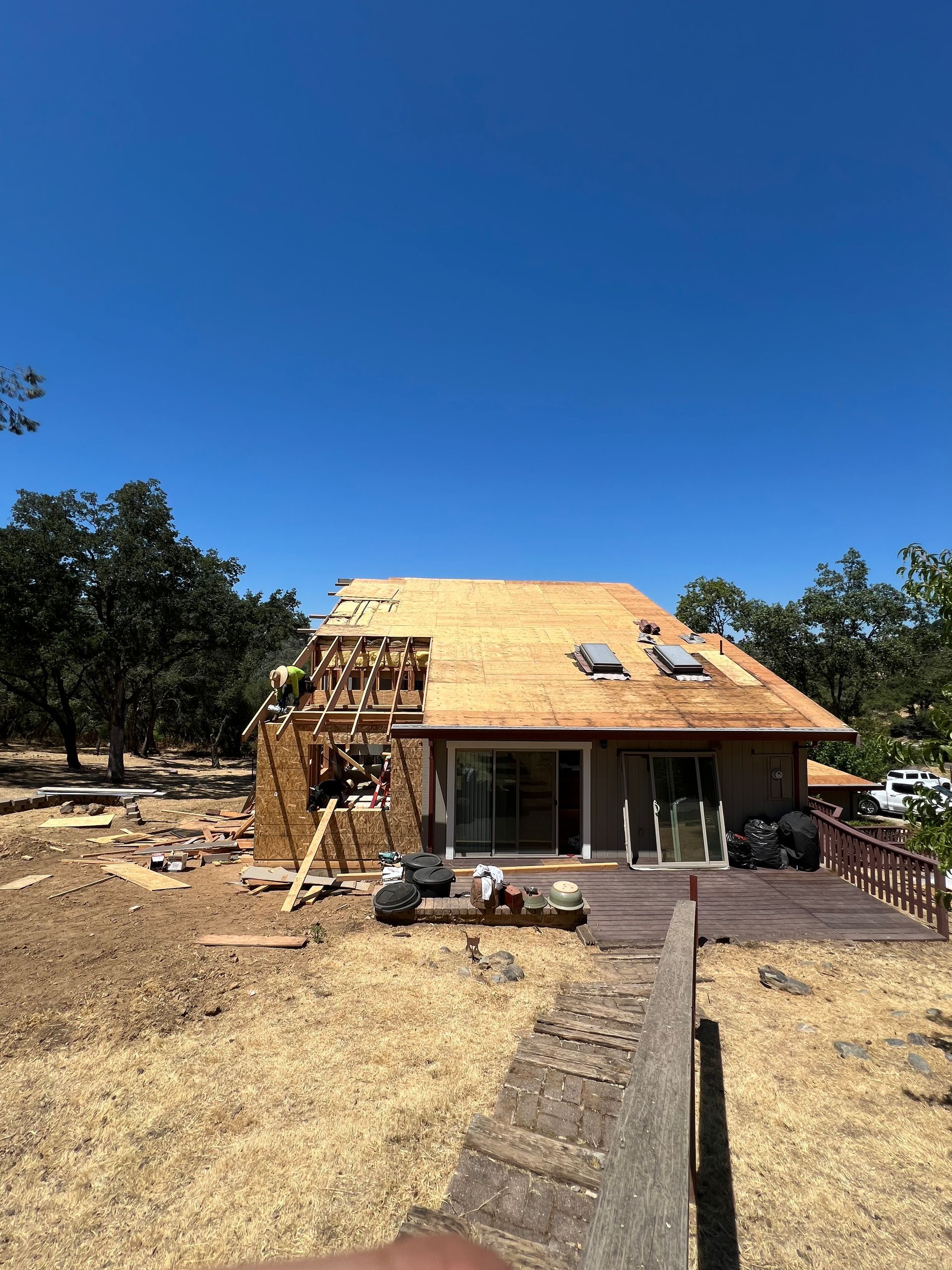 House under construction with roof partially removed, surrounded by dry grass and trees under a clear blue sky.