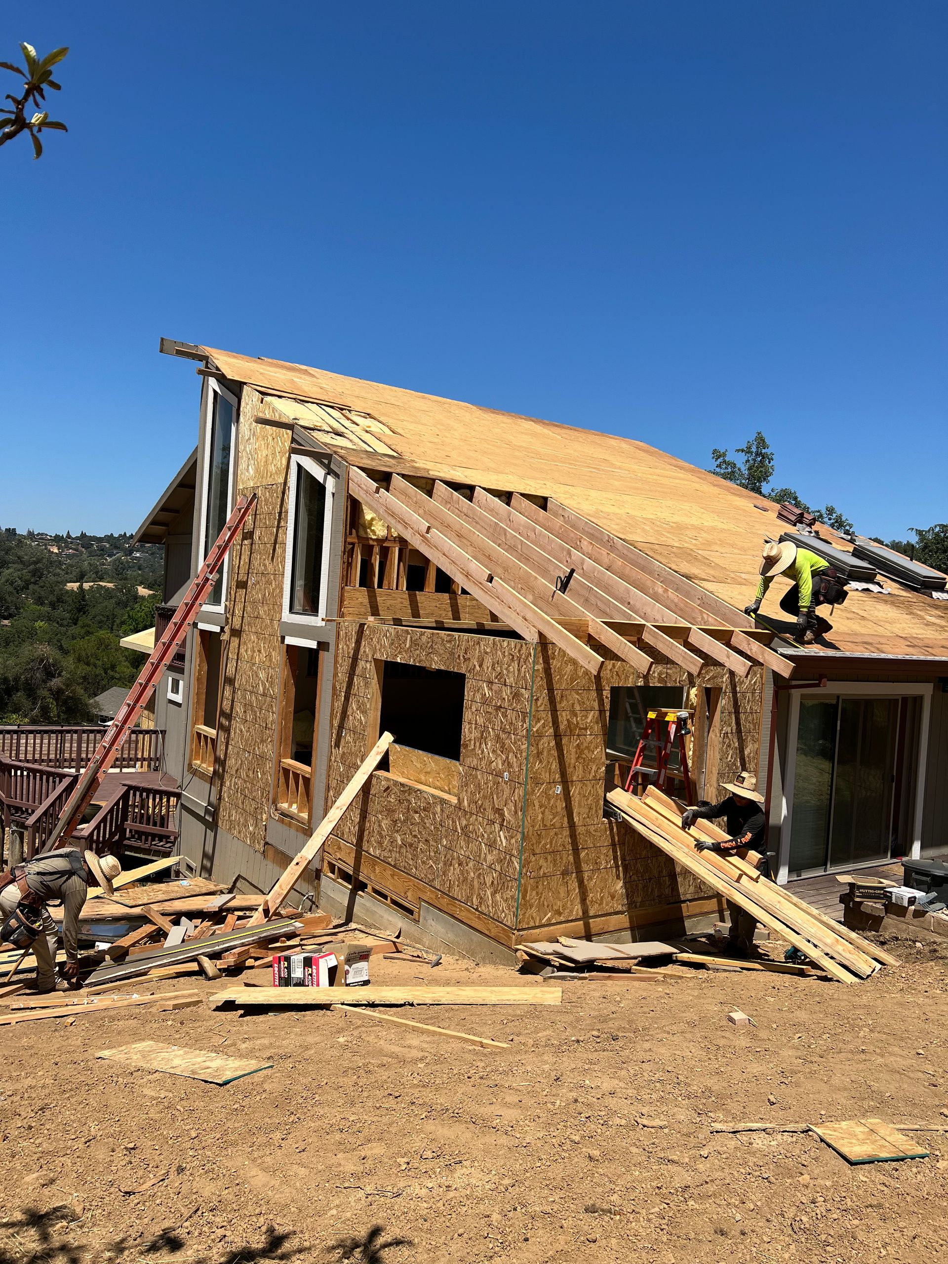 House under construction; worker on roof, wooden frame exposed, debris scattered on ground, blue sky in background.