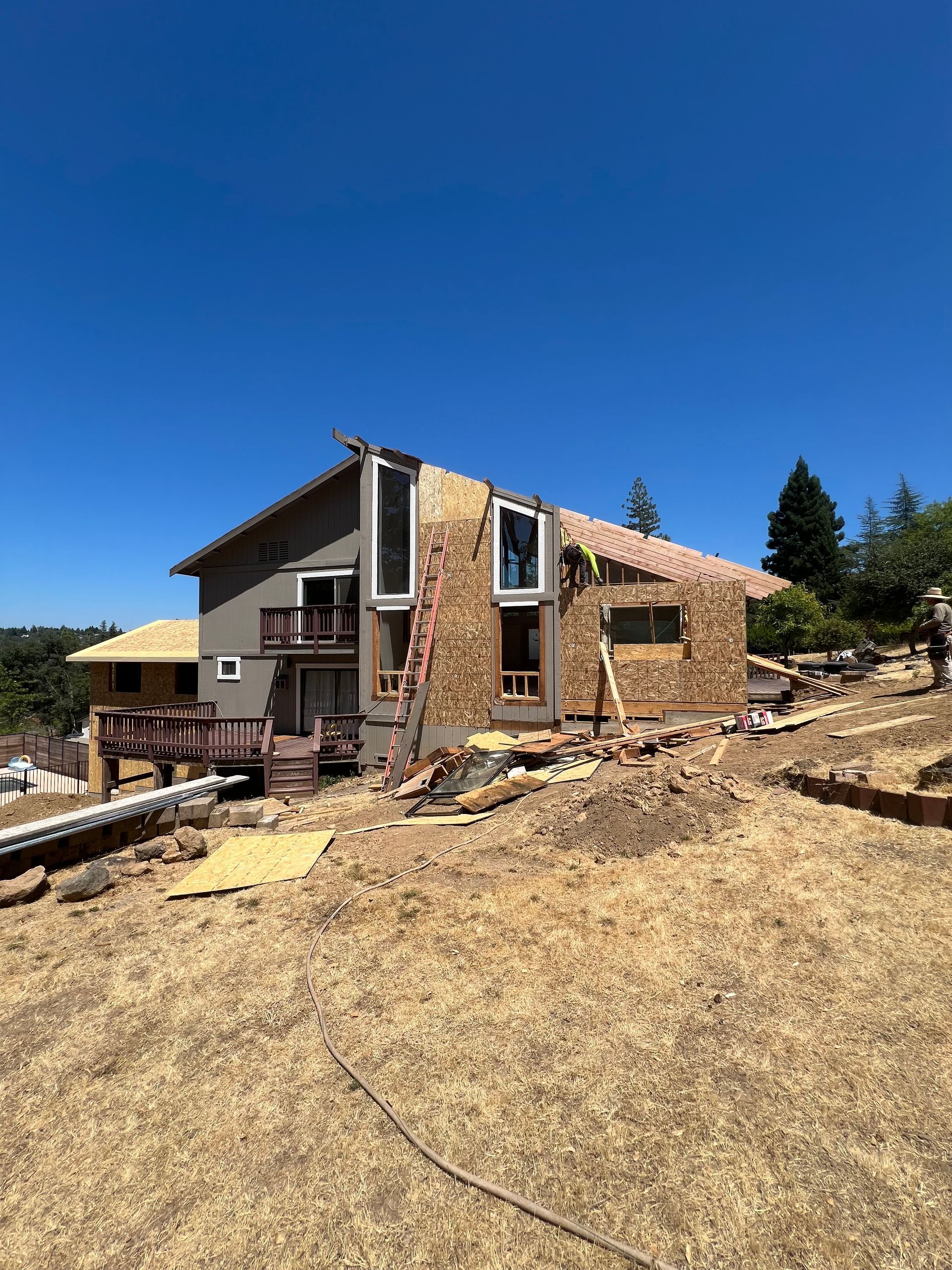 Two-story house under construction on a hillside under a clear, blue sky. Exterior walls are partially covered in wood.