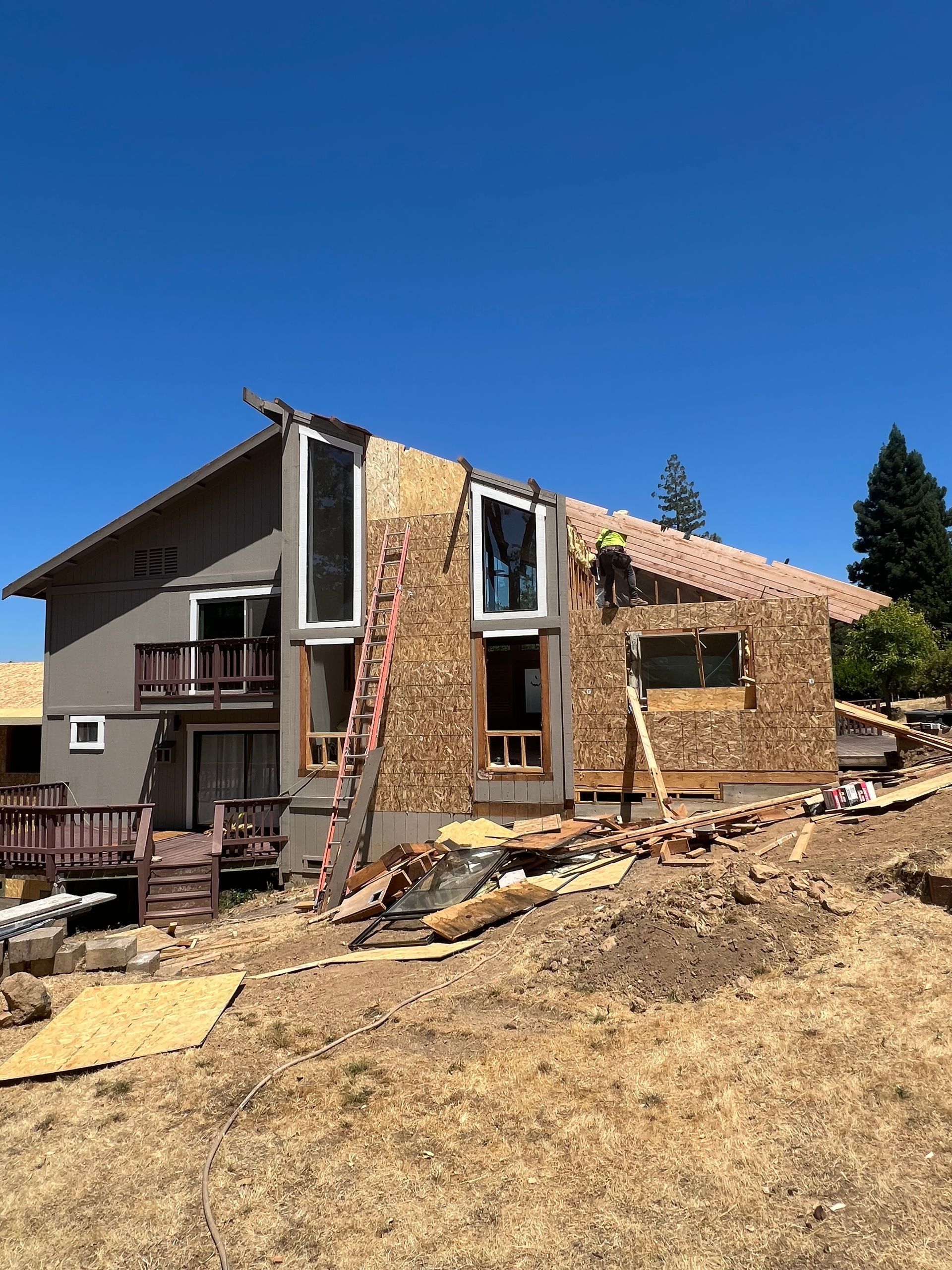 House undergoing construction; exterior walls partially covered in wood, ladder leaning against the frame. Blue sky overhead.