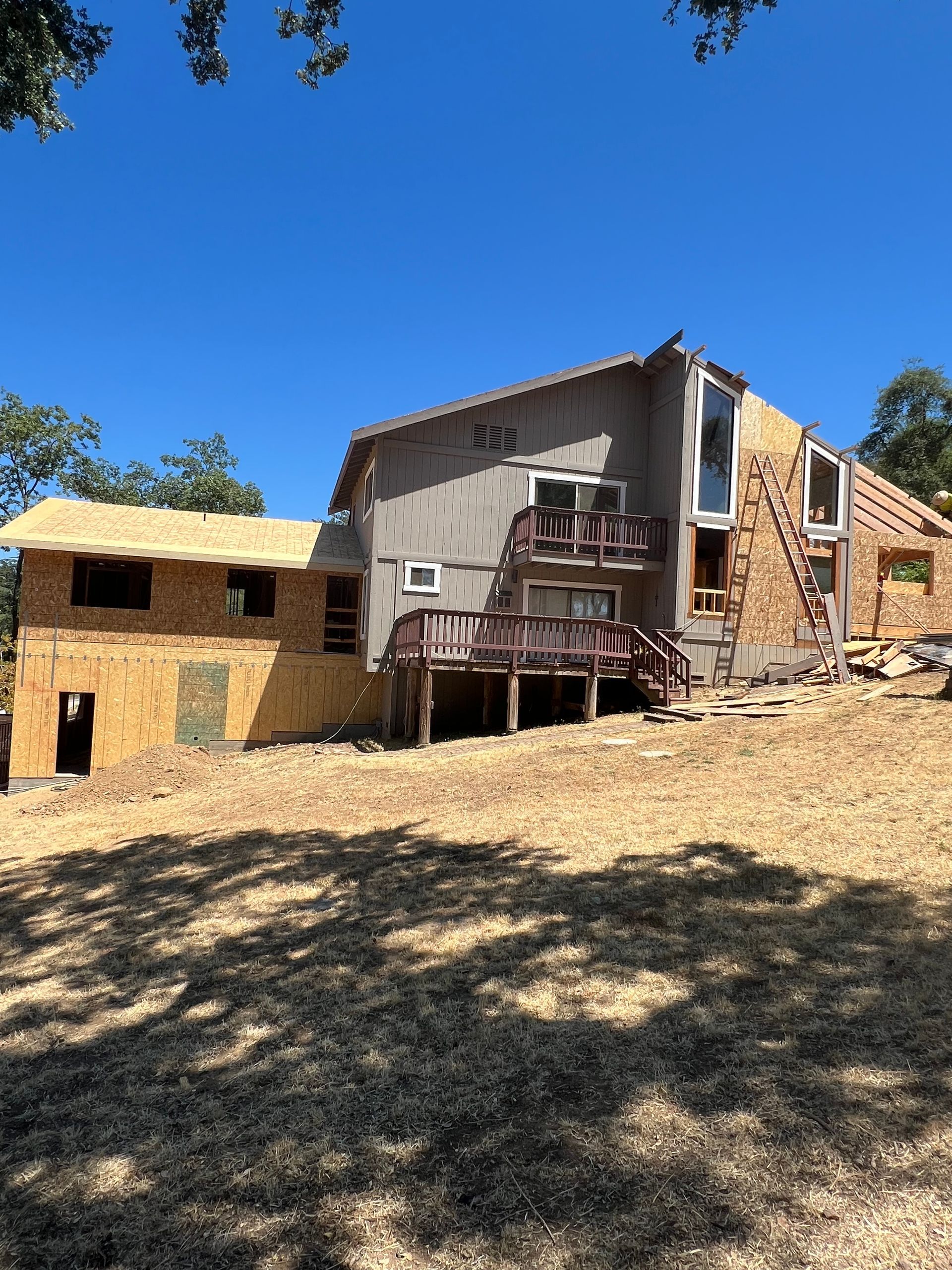 House under construction, with exposed wood framing and a completed section with stucco. Set on a grassy, sloping hillside.