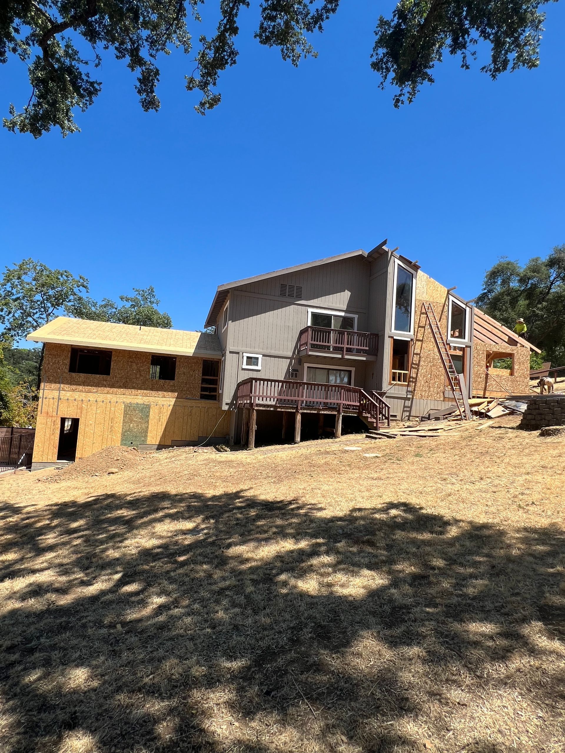 House under construction, with unfinished exterior, brown deck, and surrounding trees under a blue sky.