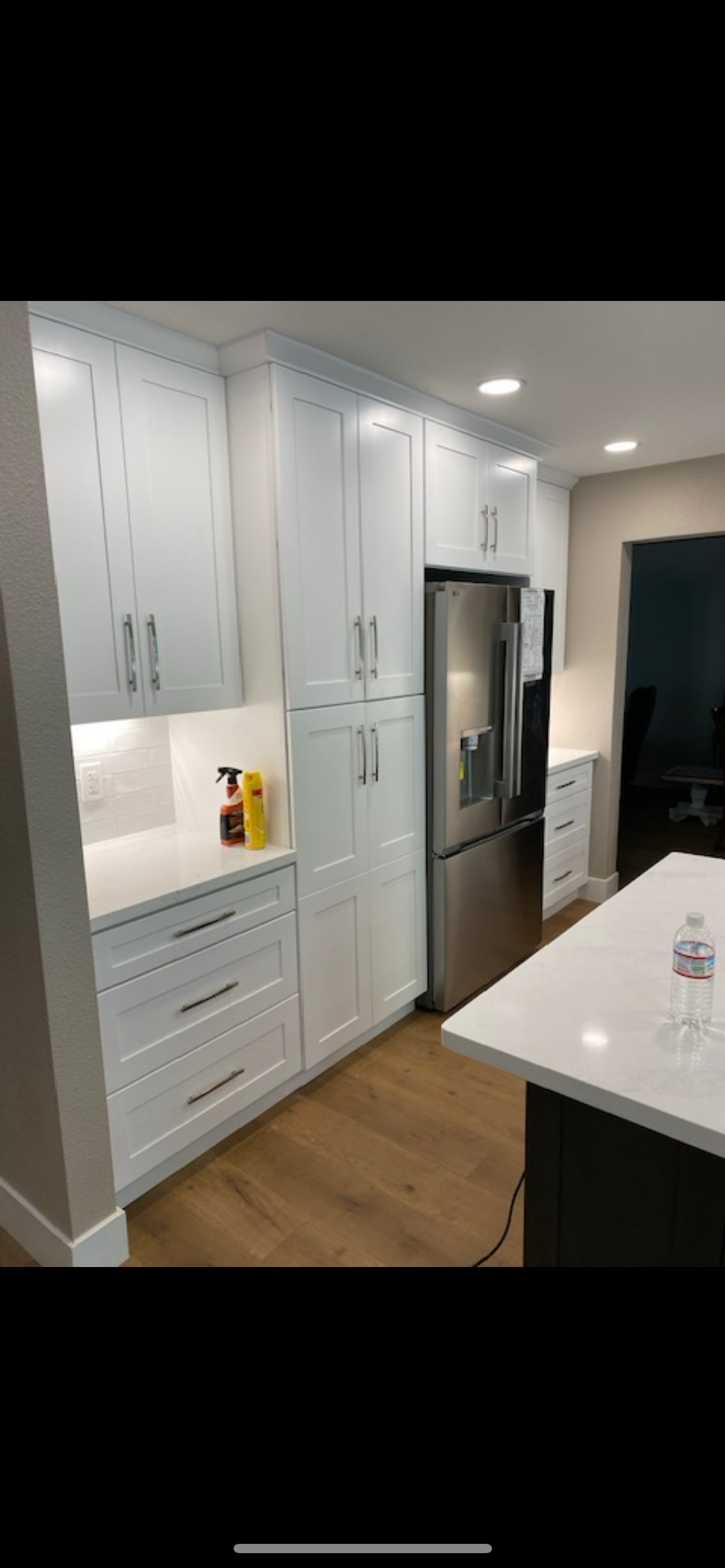 A kitchen with white cabinets, stainless steel refrigerator, and a white countertop island. Light wood flooring.