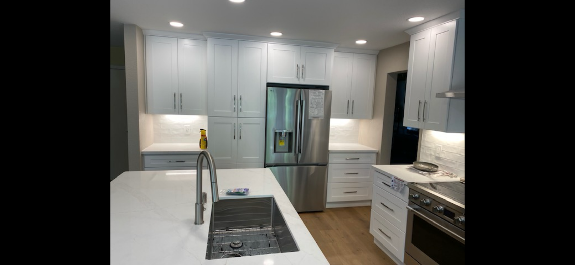 A modern kitchen with white cabinets, stainless steel appliances, a large island, and light wood floors. Recessed lighting.