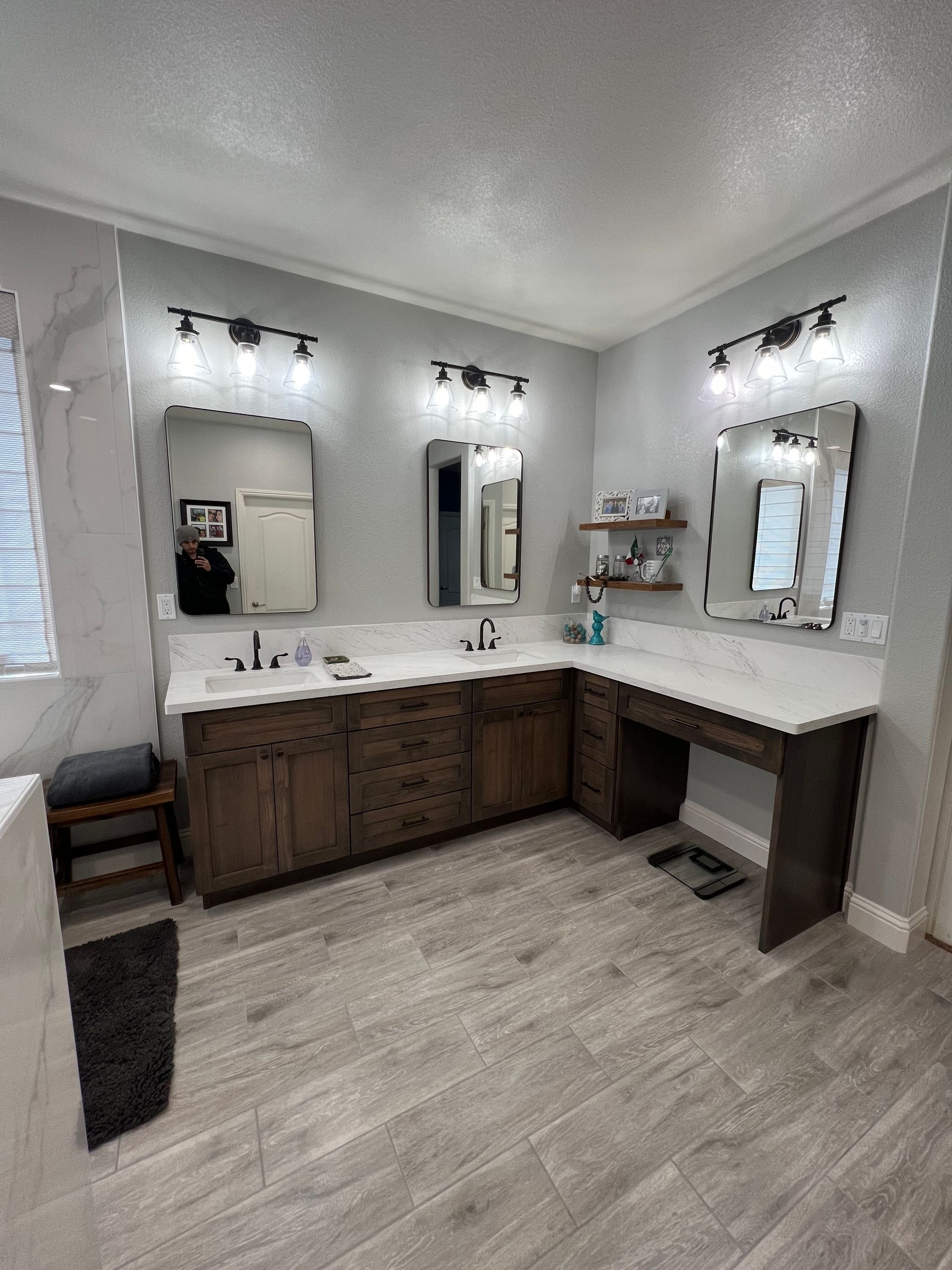 Bathroom with a corner vanity, three mirrors, and brown cabinetry. Light gray tiled floor and walls.
