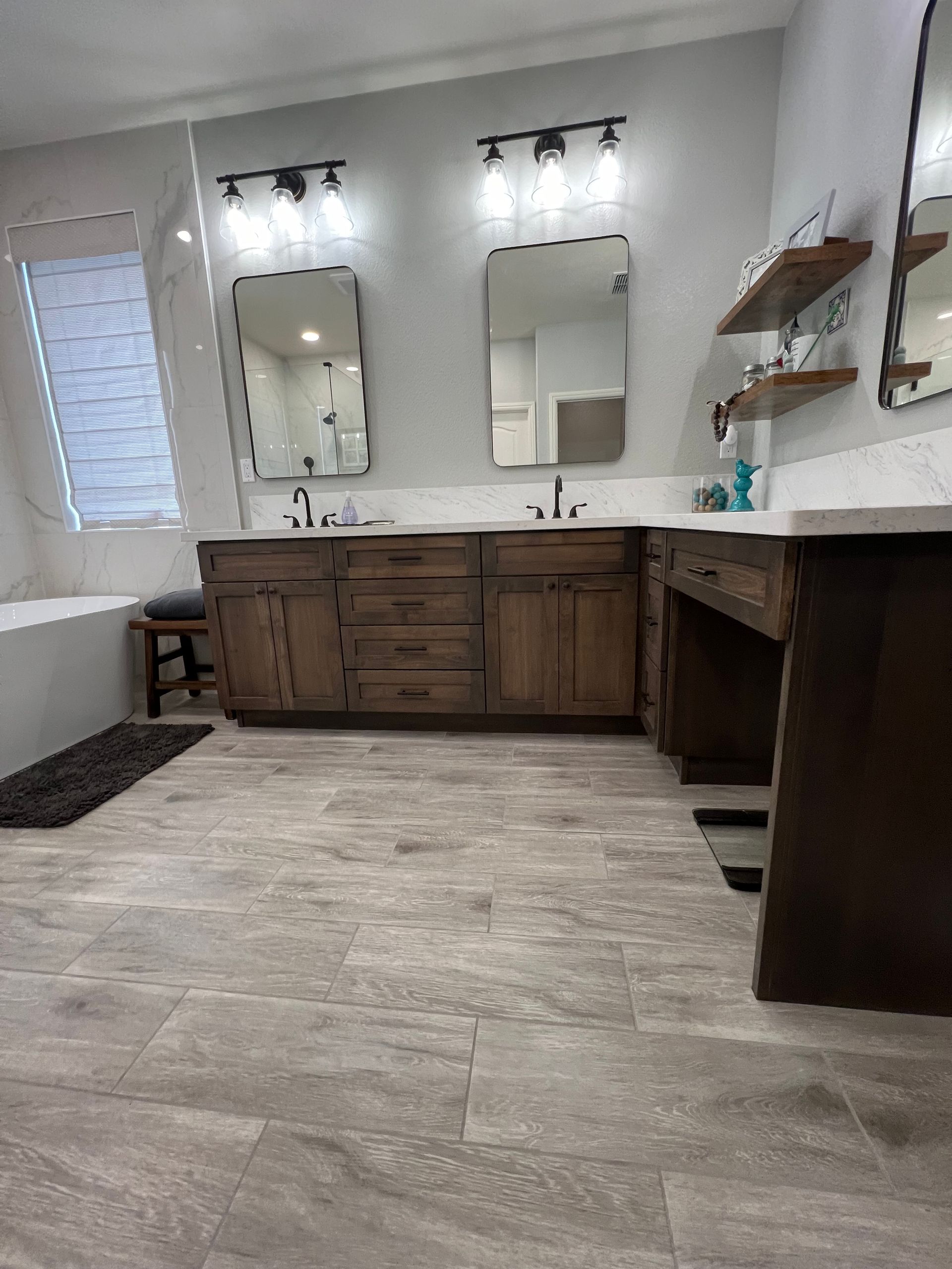 Bathroom with two mirrors above wooden vanities and light-colored tile floors. Light fixtures and shelves are visible.