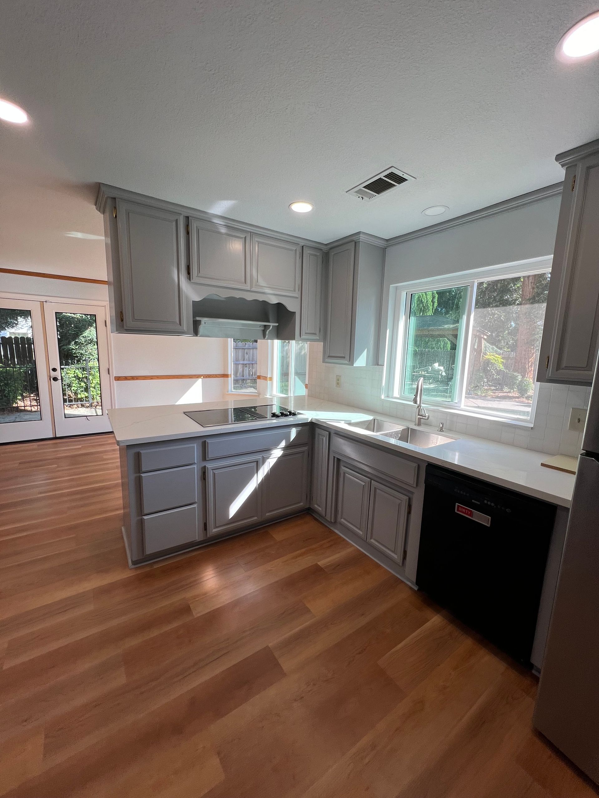 A kitchen with gray cabinets, white countertops, and wooden floors. A black dishwasher is visible.