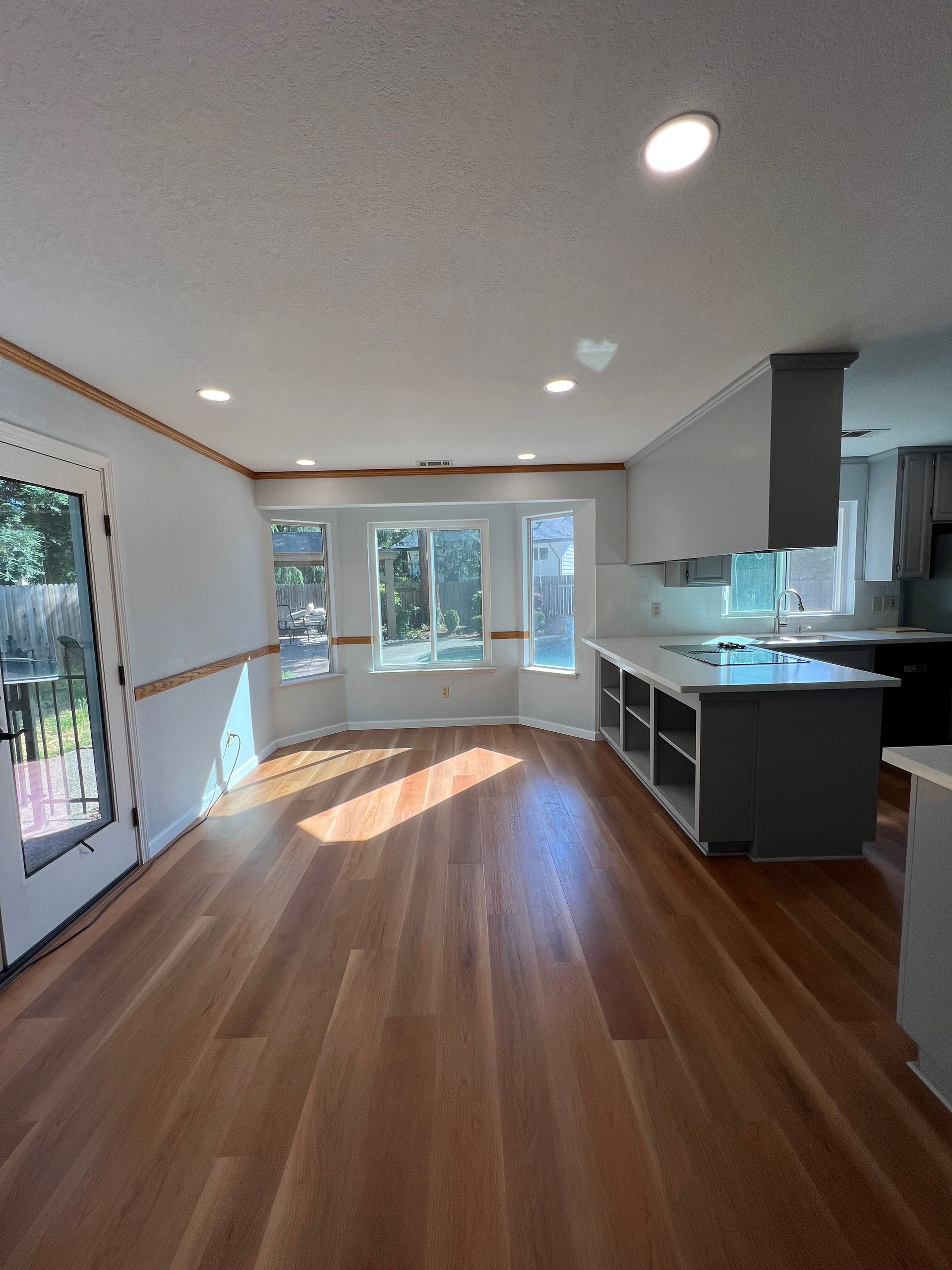 Spacious kitchen with wooden floors, a bay window, and a gray island. Bright sunlight streams in from the windows.