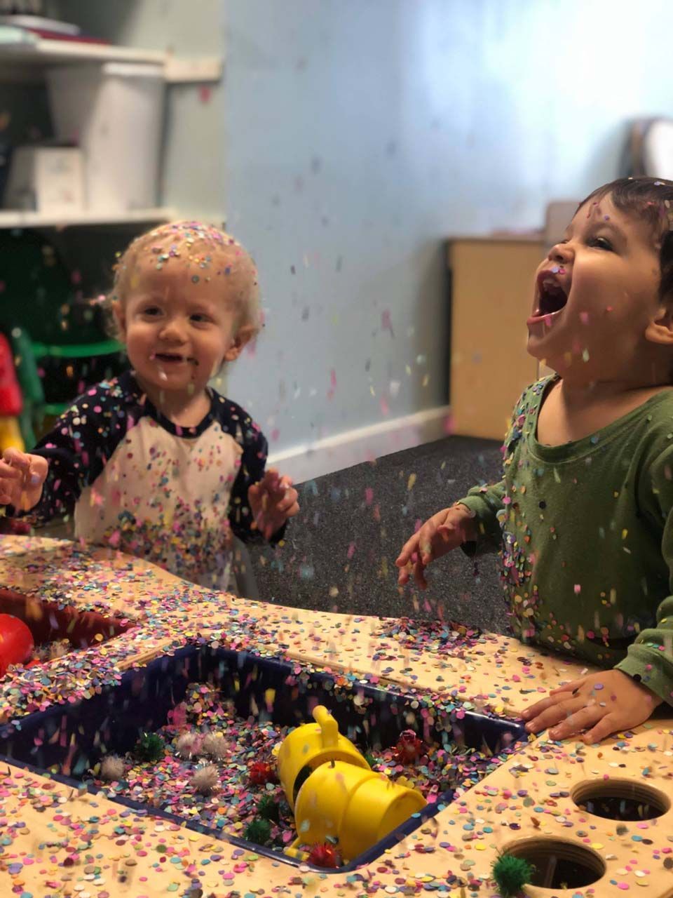 Children playing in colorful confetti at a table, laughing in a classroom or party setting