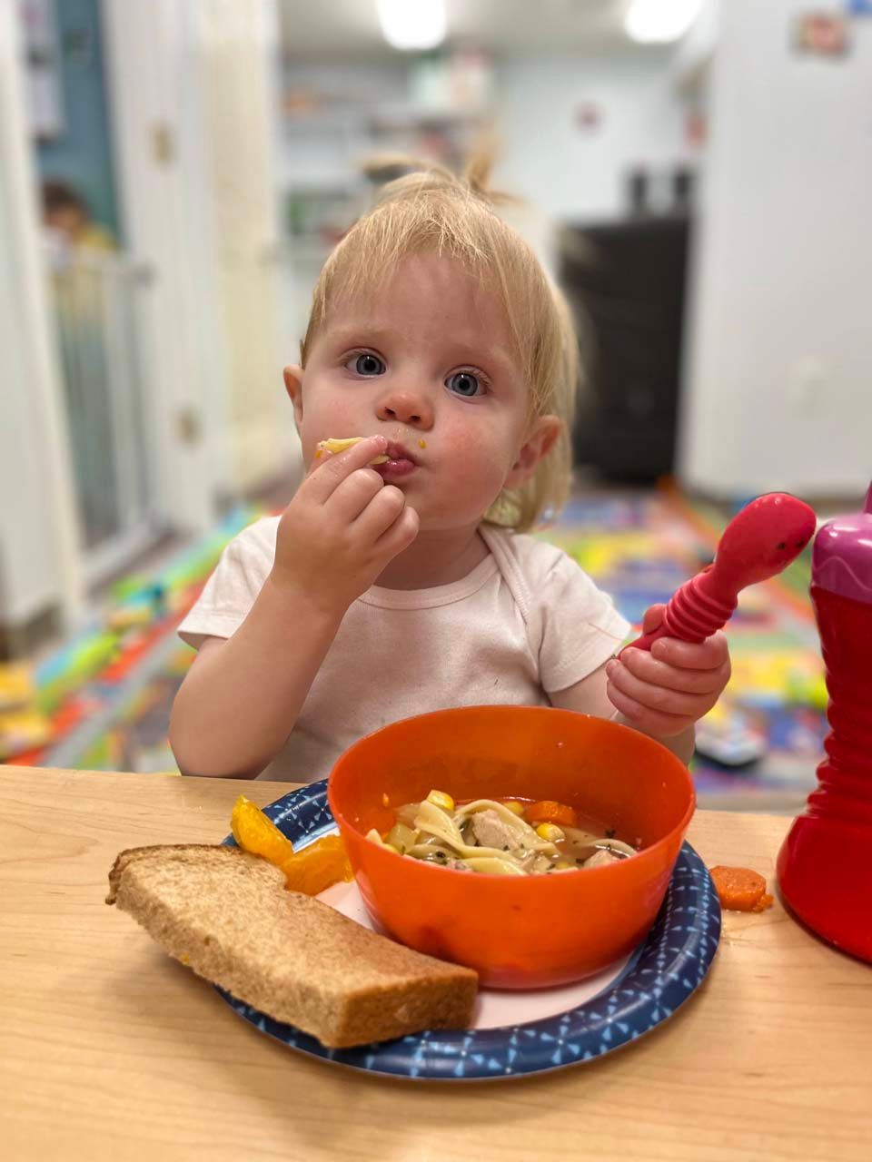 Toddler eating at a table with toast and an orange bowl, holding a pink spoon in a bright cafeteria.